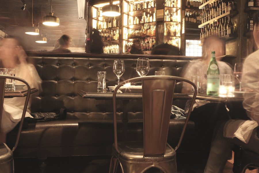 Une salle de restaurant le soir, banquette de cuir, tables dressées et bar éclairé au fond